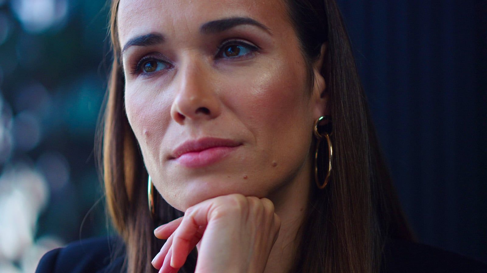 A woman with long brown hair and hoop earrings rests her chin on her hand, gazing thoughtfully into the distance—a pose psychologists might associate with mind resetting. The background is softly blurred.