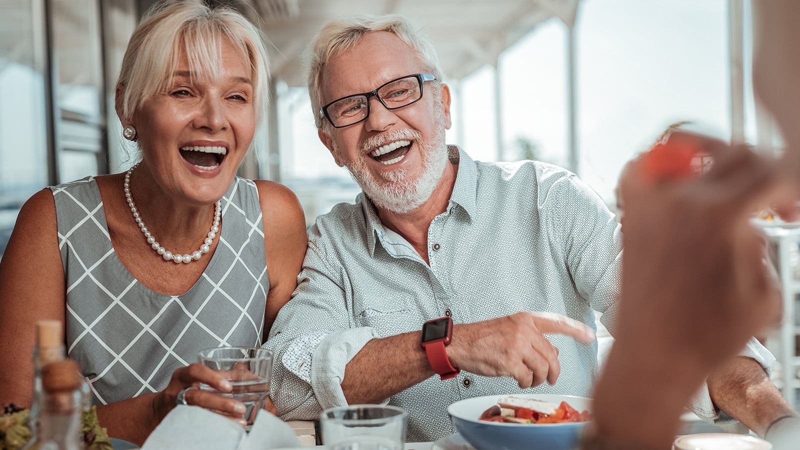 An older couple sits at a table, smiling and laughing while enjoying a meal together. The woman holds a glass of water, and the man points and laughs. Their warm connection showcases how nurturing relationships play an essential role in a successful life. The atmosphere is bright and cheerful.