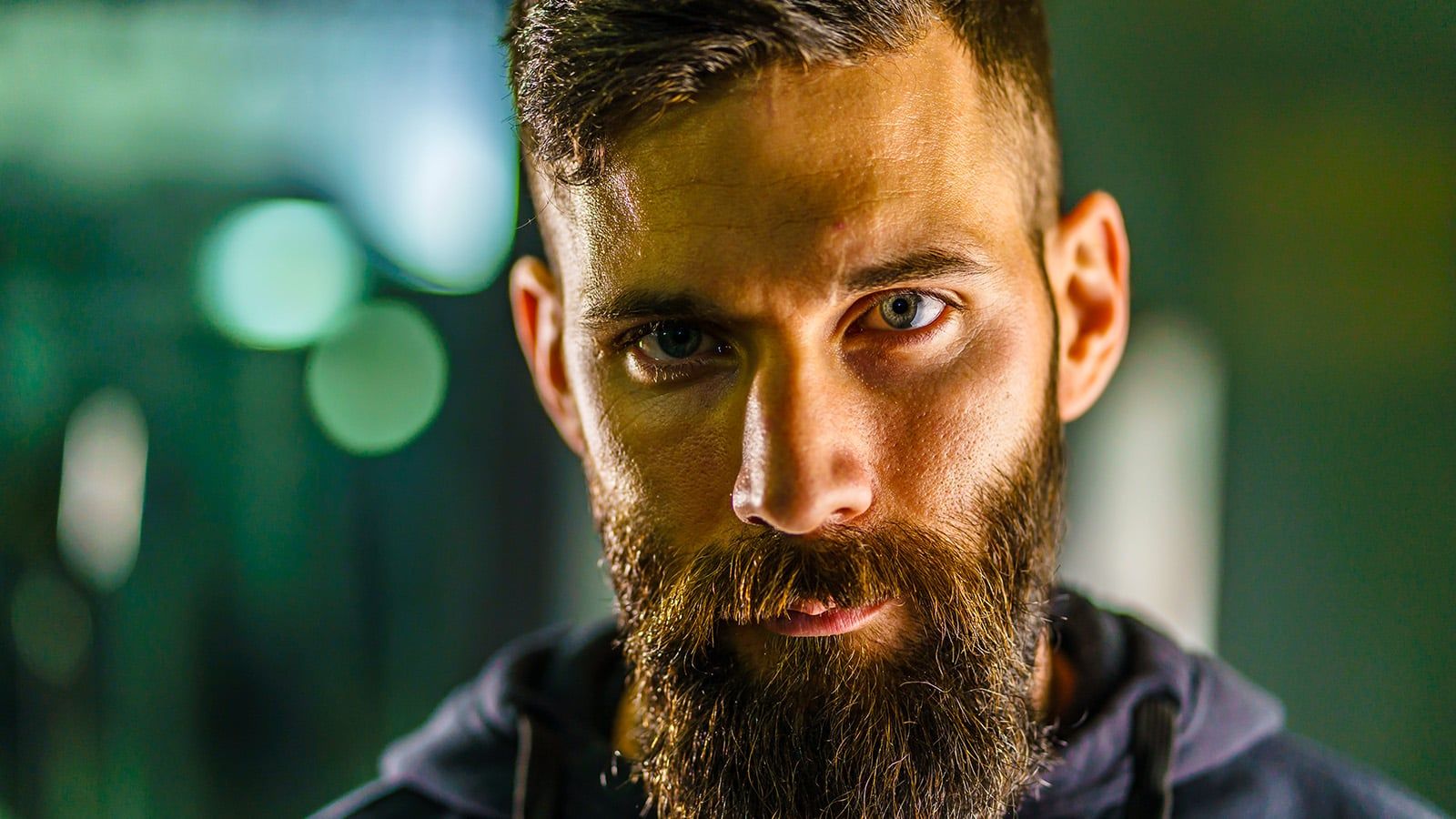 A close-up of a man with a thick beard and short, dark hair, gazing intently at the camera. He wears a dark hoodie, the blurred green and yellow lights hinting at men's feelings often hidden due to normative male alexithymia in psychology.