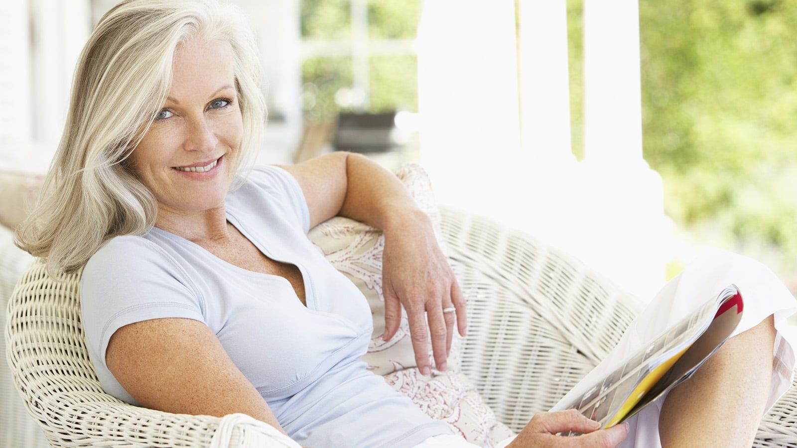 A woman with gray hair sits on a white wicker chair outdoors, smiling and holding a magazine. Radiating happiness in her 60s and 70s, she looks relaxed in her light blue top, surrounded by greenery and sunlight.