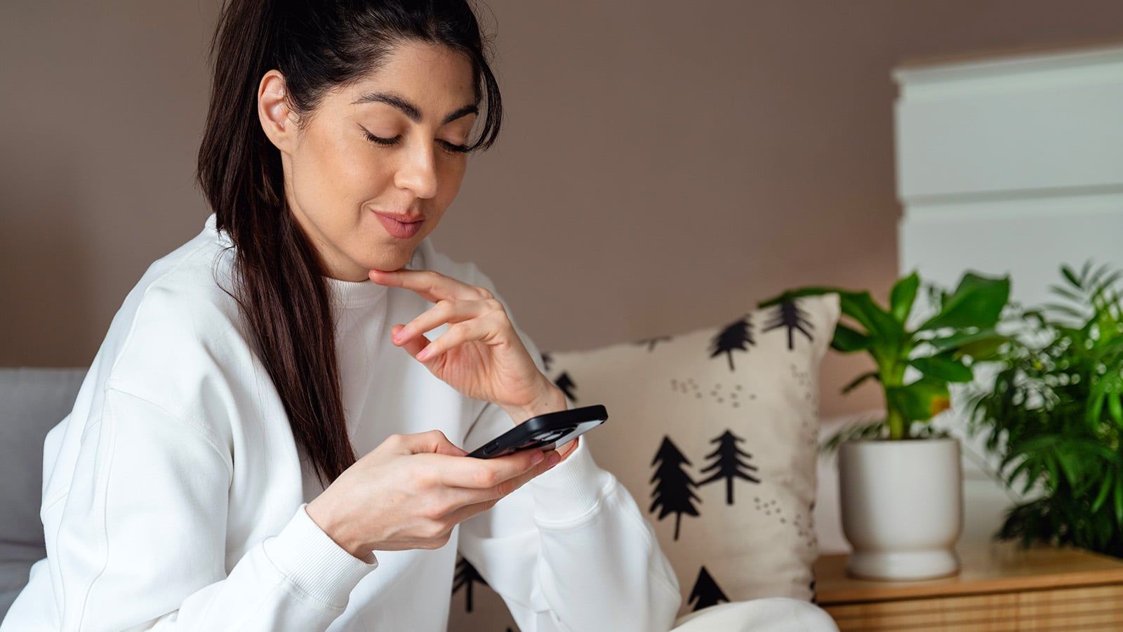 A woman with long dark hair in a ponytail, wearing a white sweatshirt, sits indoors on a couch, smiling at her smartphone—perhaps reading tips on psychology or perfectionistic self-presentation. A decorative pillow and potted plant are visible in the background.