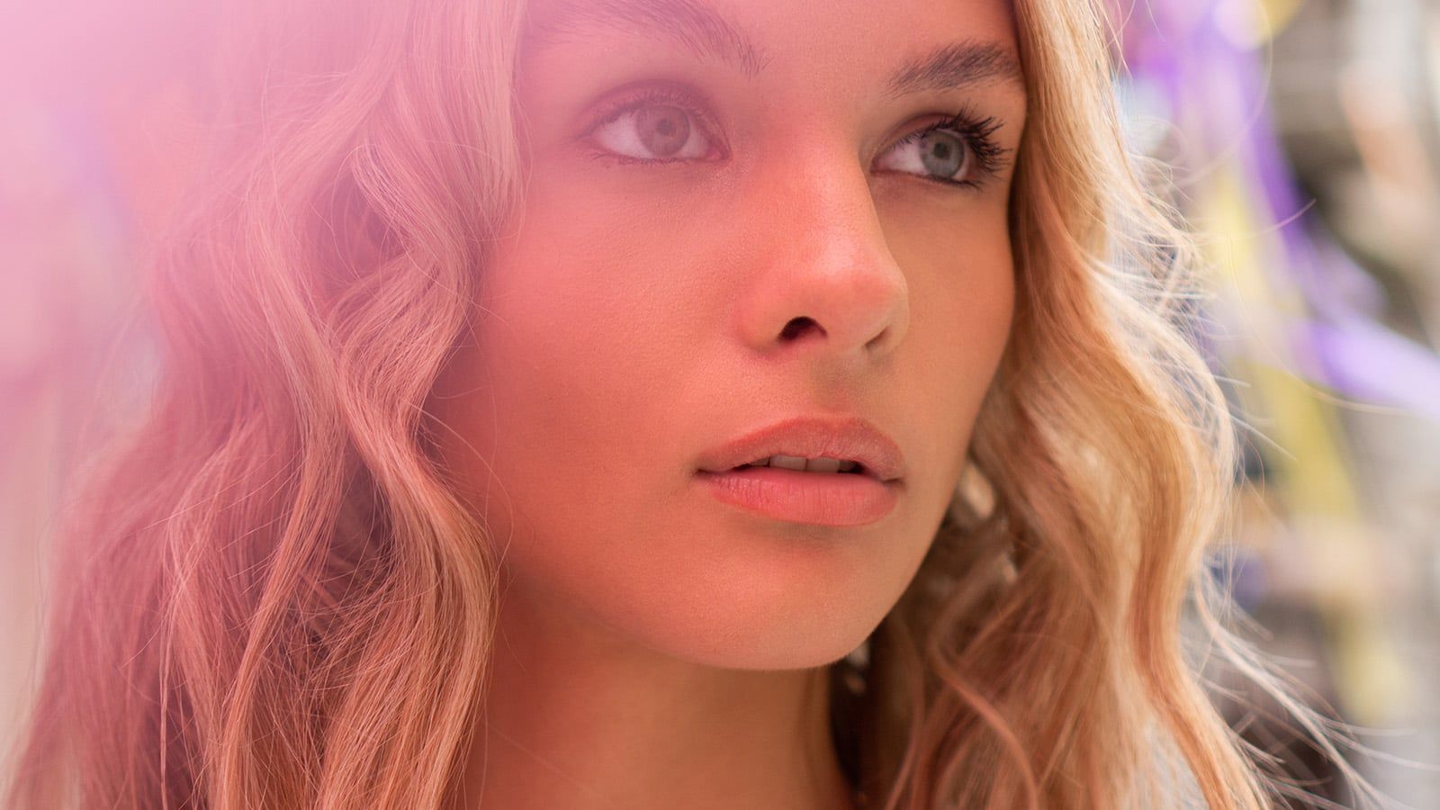 A close-up portrait of a young woman with wavy blonde hair and blue eyes, looking thoughtfully into the distance. The image has a soft pink hue and blurred background, capturing a moment of introspection about psychology and social blind spots.