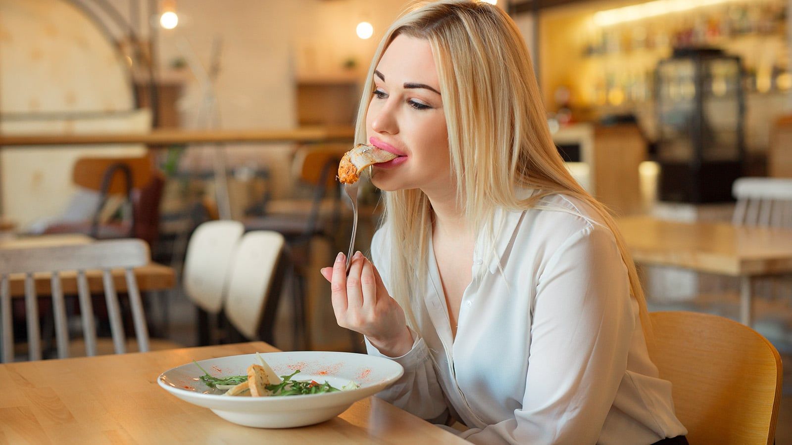 A woman with long blonde hair, wearing a white blouse, sits at a wooden table in a restaurant, enjoying a salad. The softly lit, cozy background sets the scene as she unwinds and practices healthy coping mechanisms during her meal.
