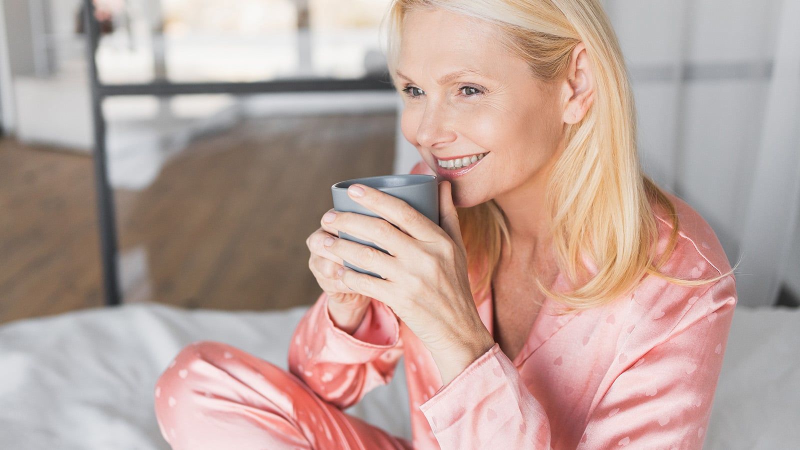 Smiling woman in pink pajamas sitting on a bed, holding a gray mug with both hands, enjoying a warm drink in a bright room—embodying the impressive life and calmest moments often celebrated in psychology for those in their seventies.