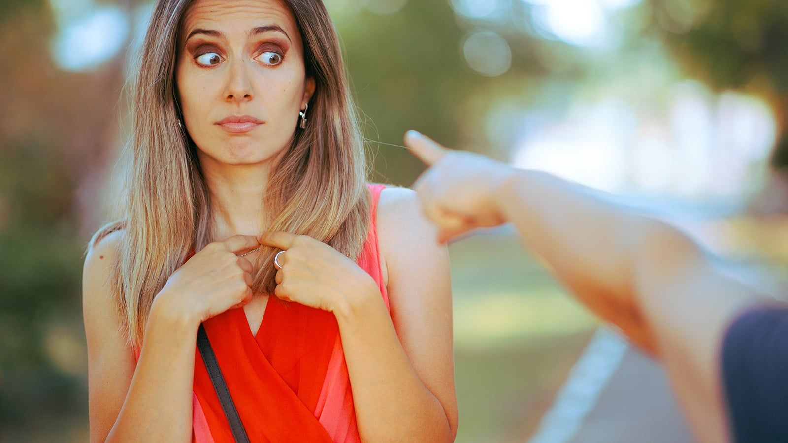 A woman with long brown hair and a red dress looks surprised and points at herself while another person’s outstretched arm points at her, capturing a moment of unexpected criticism outdoors against a blurred background.