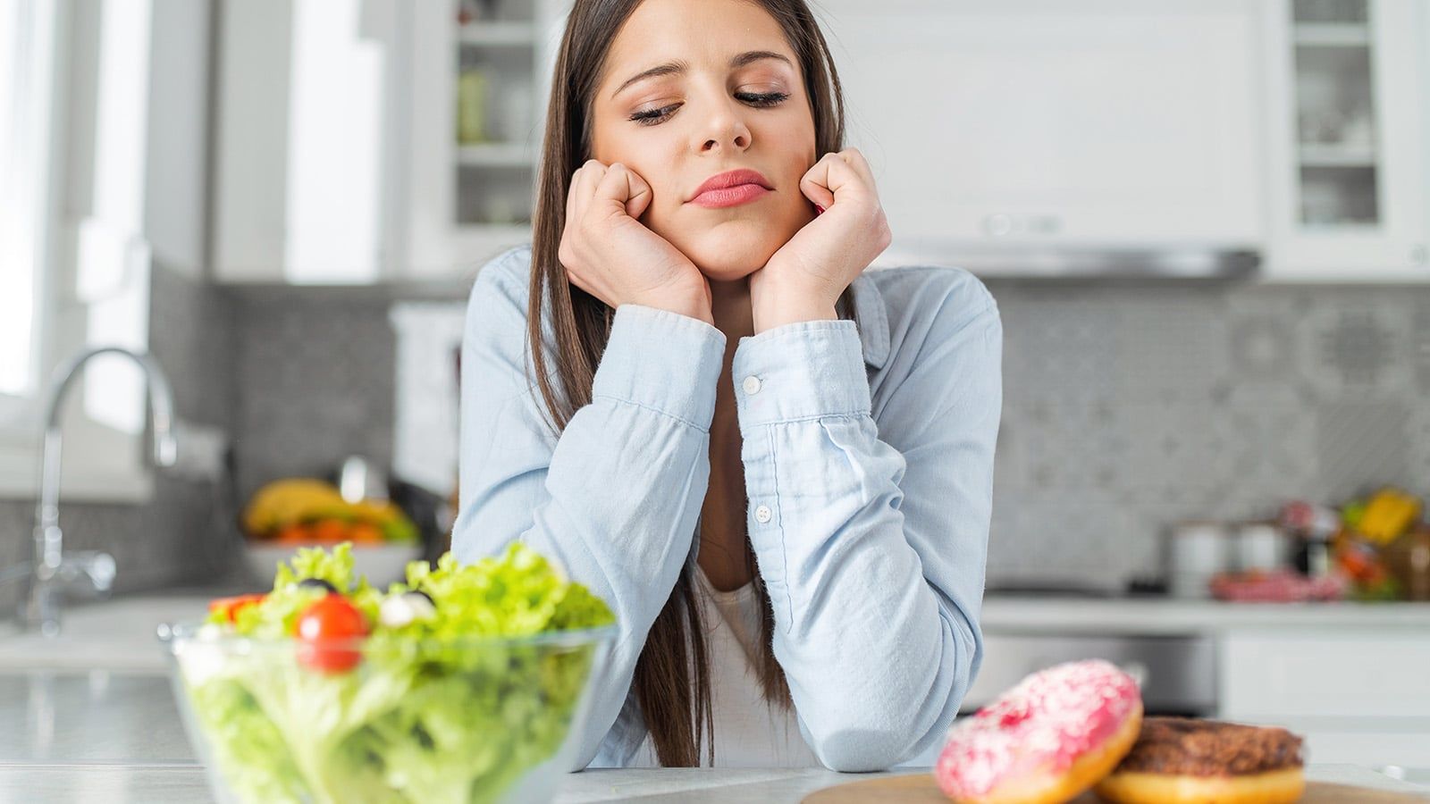 A woman sits at a kitchen counter, looking thoughtfully at a bowl of salad while donuts tempt her nearby. She appears to be exercising willpower as she faces a decision-making moment between healthy and sweet food options.