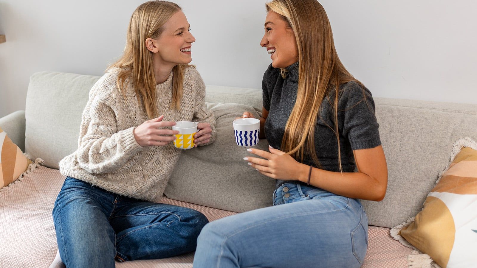 Two women with class sit on a couch, smiling and holding mugs. One wears a beige sweater and blue jeans; the other wears a gray top and blue jeans. They appear to be having a friendly conversation, each displaying unique traits that reflect their warmth and connection.