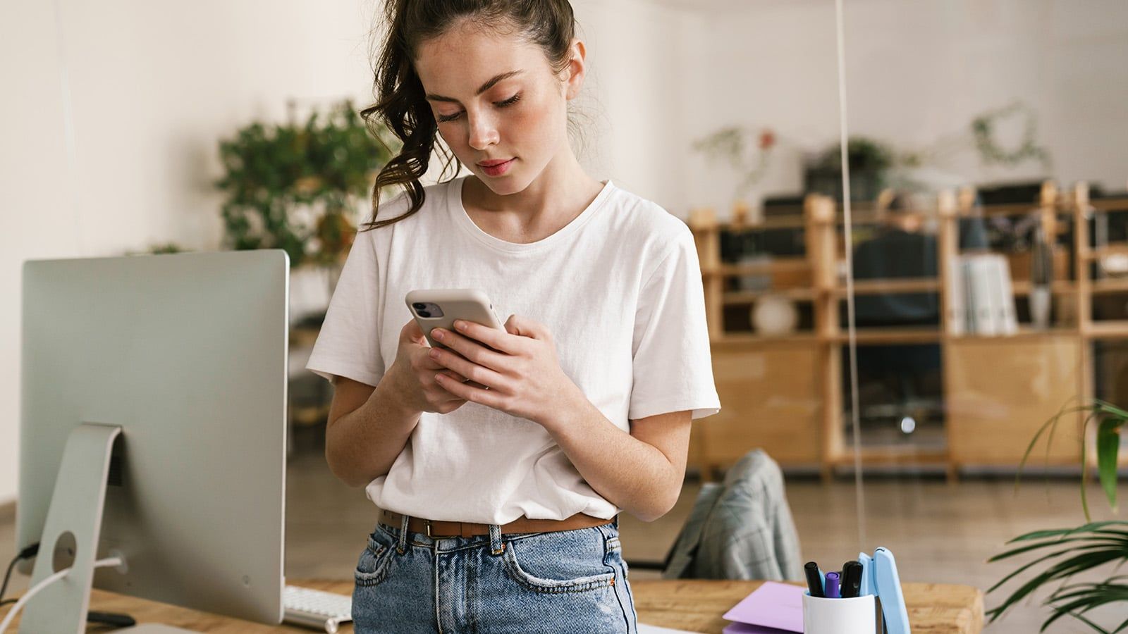 A young woman in a white t-shirt and jeans stands in an office, checking her phone. She is next to a desk with office supplies and a computer monitor, balancing productivity while surrounded by shelves and plants.