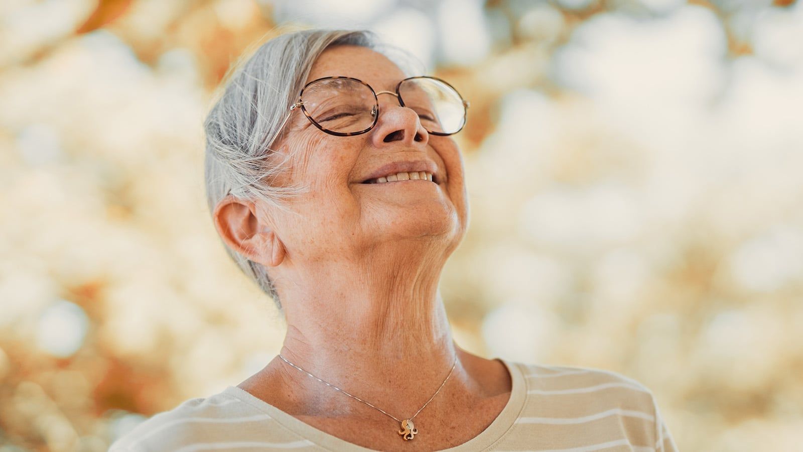 An older woman with glasses smiles peacefully with her eyes closed outdoors. She wears a striped shirt and a necklace, embodying the calm that often comes after retirement adjustment. Sunlight and blurry autumn leaves in the background create a warm setting, highlighting the anchors of comfort and tranquility that many retirees find in this new chapter of life.