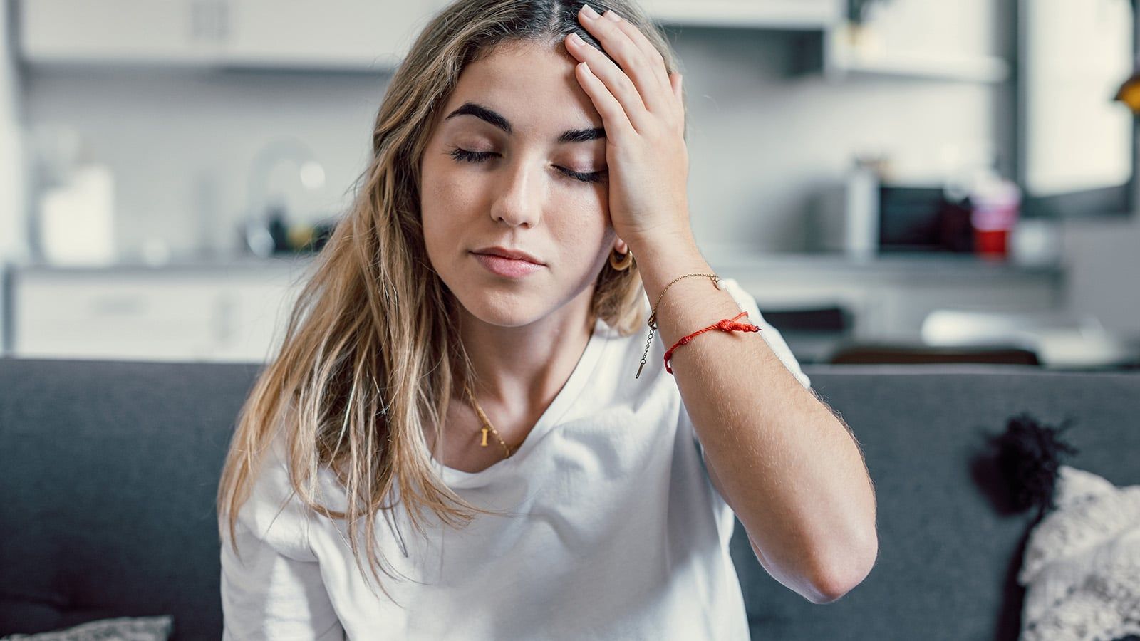 A young woman with long hair sits on a couch in a kitchen, eyes closed, and holds her hand to her forehead, appearing tired from hard work. She is wearing a white shirt and gold jewelry.