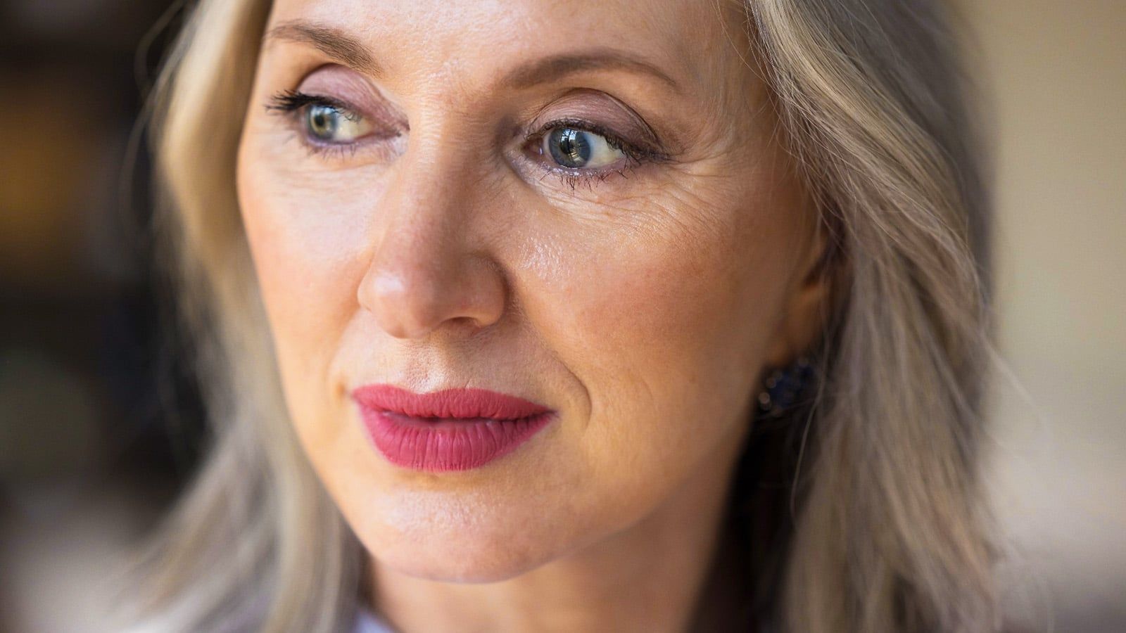 A middle-aged woman with light skin and blonde hair looks thoughtfully to the side, her calm expression perhaps hinting at retirement loneliness or the effects of winter depression. She has pink lipstick, subtle eye makeup, and wears a gentle demeanor. The background is softly blurred, emphasizing her quiet contemplation in a moment touched by social isolation.