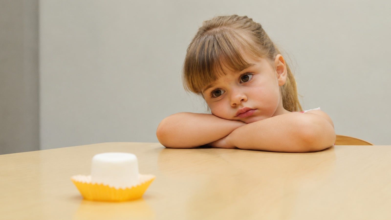 A young girl with blonde hair rests her chin on her crossed arms, gazing thoughtfully at a single marshmallow in a yellow wrapper—a quiet moment of willpower as she faces her own marshmallow test.