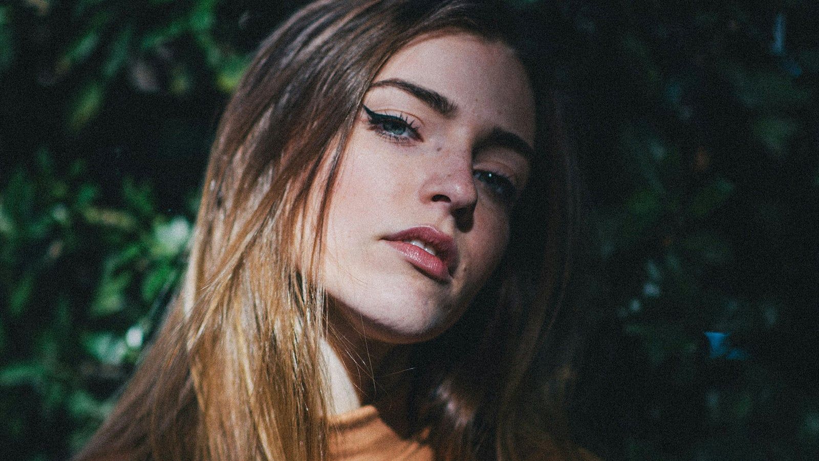 A young woman with long, light brown hair and blue eyes looks slightly upward, standing against a background of dark green foliage. She wears a neutral expression and a light brown top, as if lost in thought about beliefs and actions. Sunlight highlights her face.