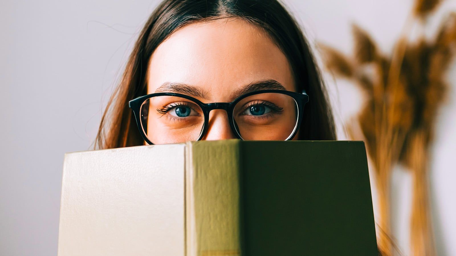 A person with long brown hair and glasses—displaying signs of high IQ—looks over the top of an olive green book, partially covering their face. The background is softly blurred with dried plants visible.
