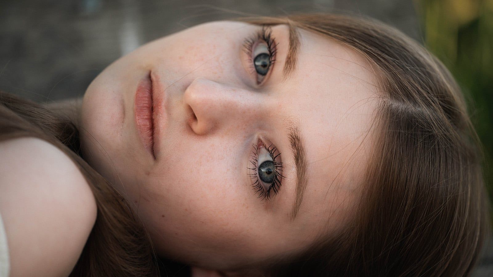 A young person with long brown hair lies on their side, gazing directly at the camera with blue eyes. The close-up highlights their clear skin, natural makeup, and serene expression—yet there’s a quiet intensity that hints they may have grown up walking on eggshells, carefully choosing their phrases even in moments of stillness.