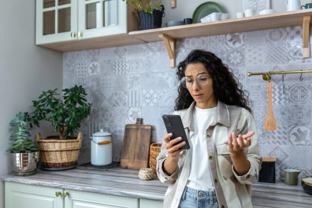 A sad young hispanic woman in the kitchen, pressing her phone and surprised by what she sees on her phone