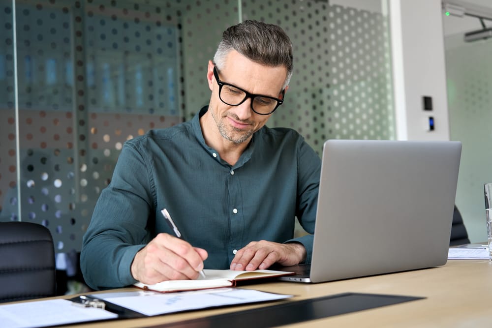 A busy middle age mature man writing in his notepad with his laptop in front of him