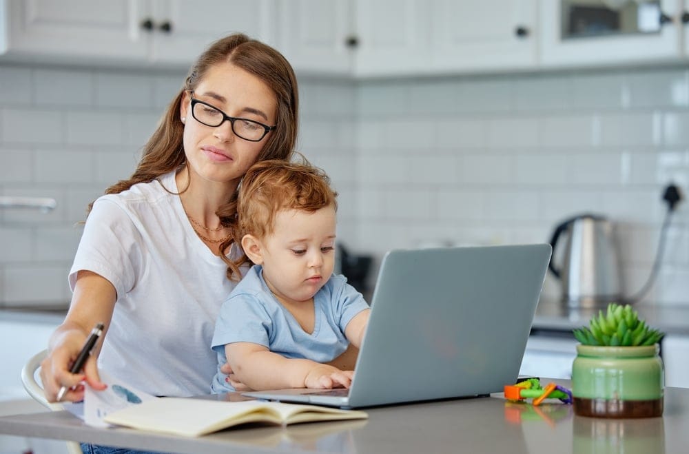 A mother sitting in front of her laptop, her young son is sitting on her lap and he is using the laptop, his mother is reading a book beside the laptop