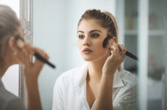 Closeup shot of attractive mid 20's blond woman putting on some makeup in the morning before going out. She's standing in front of big bathroom mirror, rear view.