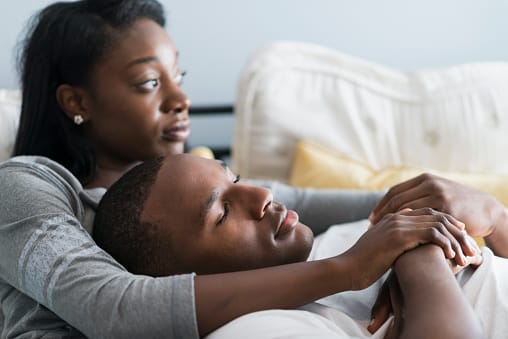 A young African American couple lying bed. The focus is on the man who is asleep with his head on his girlfriend's chest. She is awake, looking away with a serious expression.