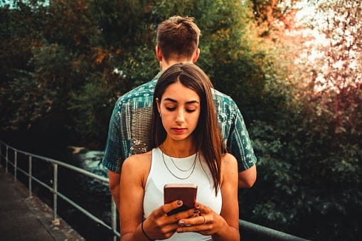 Young woman checking messages on mobile phone while standing back to back outdoors with her boyfriend. Reading concentrated the new messages and social media posts on her smart phone. Teenage Youth Culture Smart Phone Social Media Addiction Lifestyle Concept.