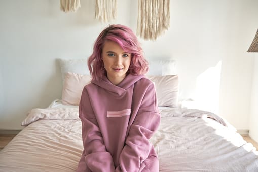 Smiling hipster gen z teen girl with pink hair wear hoodie sit on bed, portrait.