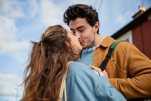 A low angle close up view of a young couple affectionately embracing in the street in Whitley Bay. They are standing still and kissing with their eyes closed.