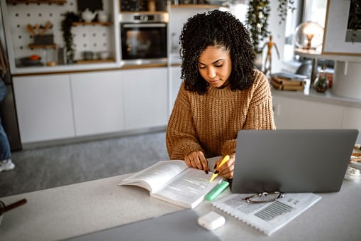 Young pretty student woman with curly hair using laptop for studying while her roommates standing in the background in their campus apartment.
