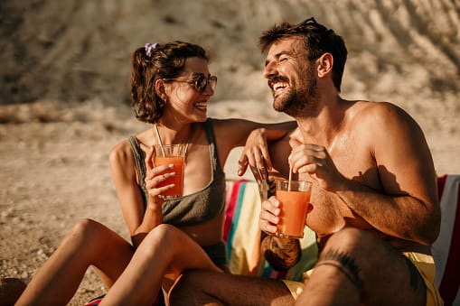 Photo of a cute brunet with sunglasses, and her bearded friend, enjoying sunbathing by the sea/lake, in their swimming suits, drinking a freshly squeezed juice/cocktails, facing each other while enjoying conversation and laughter. Waiting for the rest to come