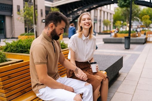 Happy young couple sitting on bench and drinking takeaway coffee enjoying time together outdoors on summer. Attractive young woman and bearded man having carefree talk sitting on bench holding hands.