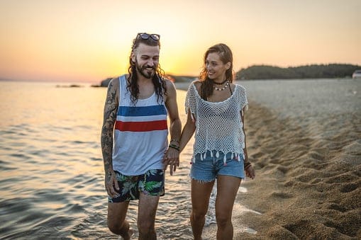 Photo of happy young couple walking on the beach holding hands. Young man and woman enjoying beach holiday at sunset.