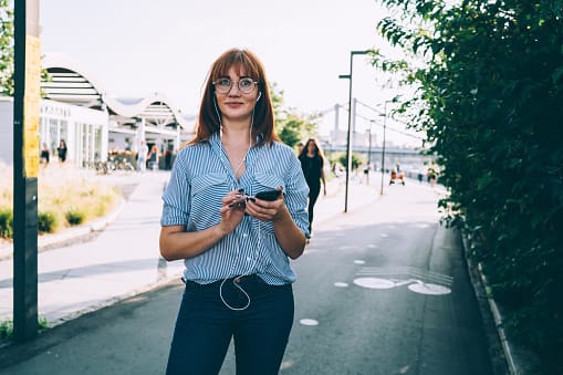 Full length portrait of beautiful caucasian woman in eyewear enjoying free time outdoors using accessory and mobile phone for listening audiobook, cheerful millennial hipster girl melloman outdoors