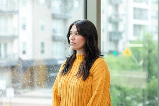 A sad young woman daydreams while looking through a window on a rainy, gloomy day.
