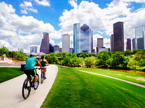 Young Man and Woman Riding Bikes on Paved Trail in Houston's Buffalo Bayou Park (view of river and skyline of downtown Houston) - Houston, Texas, USA