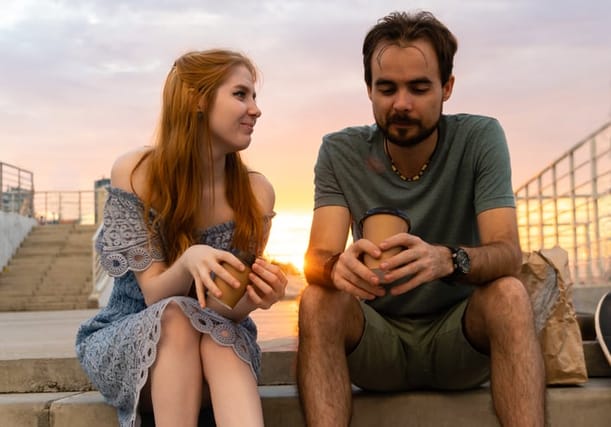 Positive young couple sitting on stone stairs with cups of coffee in hands. Smiling woman looking at boyfriend against sunset sky