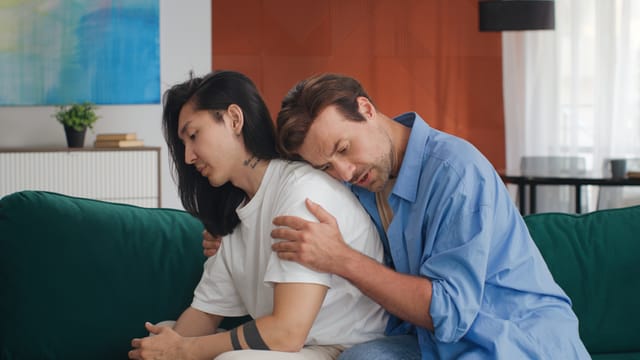 Young man hug and comfort upset gay partner sitting on couch at home. Homosexual boyfriend console and support depressed asian guy
