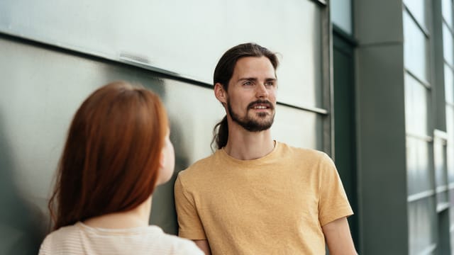 Attractive bearded young man looking aside gazing ahead with a serious expression in an over the shoulder view past a young redhead woman against a grey exterior wall