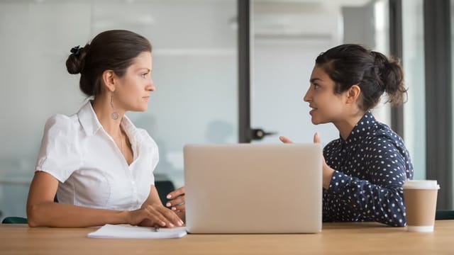 Enthusiastic young indian employee sharing new project ideas with serious female boss. Diverse business woman team discussing working moments at office. HR manager listening to interns experience