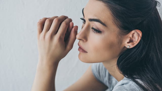 A young woman with long dark hair looks thoughtful and concerned, resting her hands near her face while gazing to the side against a plain white background.