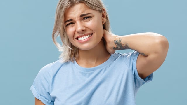 A young woman with short blonde hair wearing a light blue t-shirt looks uncomfortable and holds her neck in pain, grimacing against a plain blue background.