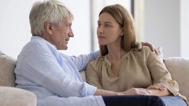 An older boomer father and a younger woman sit close together on a couch, facing each other and having a serious conversation about rent. The man’s arm is around the woman’s shoulders, and they both appear thoughtful.