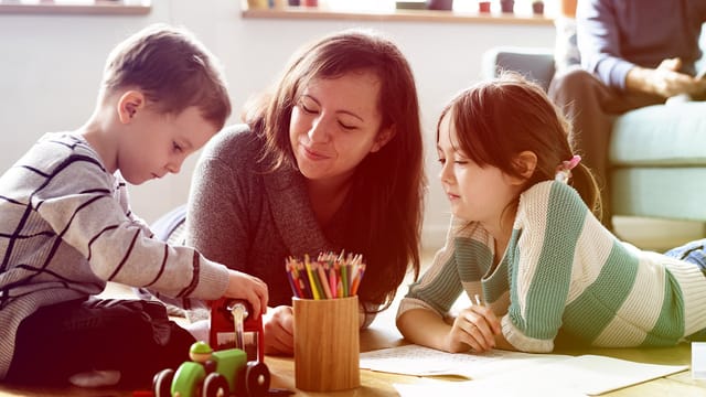 A woman—possibly a biological parent or stepparent—and two children lie on the floor, drawing with colored pencils. The boy plays with a toy tractor as the girl smiles. Papers and a container of pencils are scattered before them, capturing the joy of loving a child.
