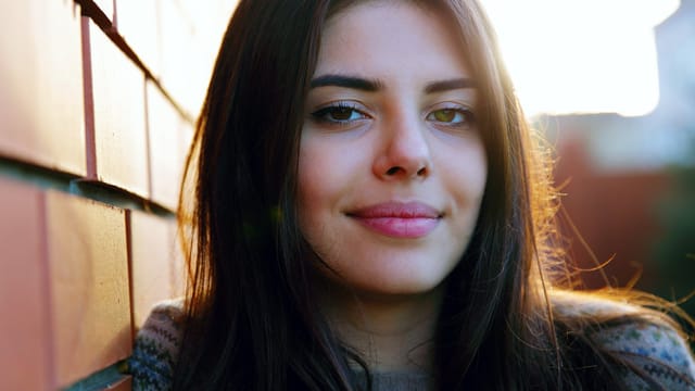 A young woman with long brown hair stands outdoors, leaning against a brick wall, softly smiling at the camera with sunlight illuminating her face—a quiet moment of self-care in today's busy world.