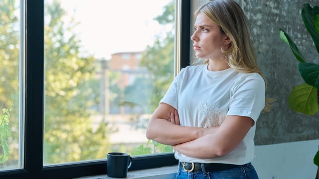 A young woman with long blonde hair, wearing a white t-shirt and jeans, stands indoors with arms crossed, looking thoughtfully out a large window—an air of anticipatory nostalgia in her gaze. A black mug sits on the windowsill beside her.