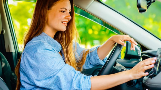 A smiling woman with long brown hair sits in the driver’s seat of a car, wearing a blue shirt and adjusting the touchscreen display, enjoying being alone in the car as green trees pass by outside.