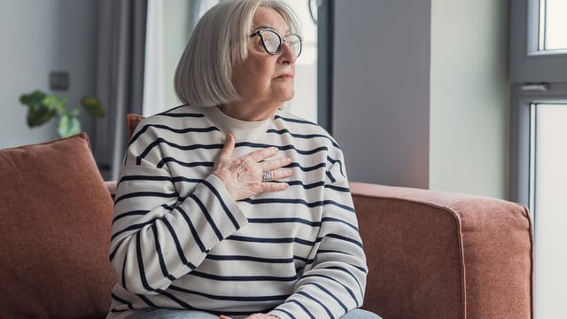 An elderly woman with gray hair and glasses sits on a sofa, wearing a striped sweater. She places a hand on her chest and looks thoughtfully out the window, reflecting on loneliness as gentle natural light fills the room.