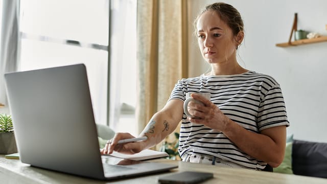 A woman in a striped shirt sits at a table, focused on her laptop with a mug in hand. Surrounded by a notebook, pen, and phone, she reflects the challenges of work-life balance in today’s digital workplace.