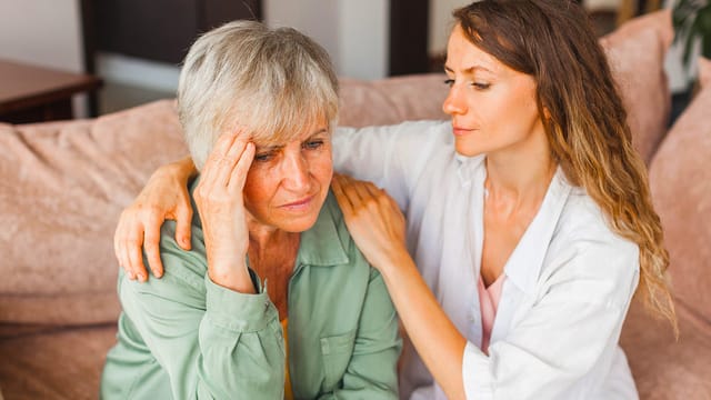 A younger woman sits beside an older woman on a couch, comforting her through grief. The older woman, mourning and distressed, holds her head with one hand while the younger gently embraces her shoulder.