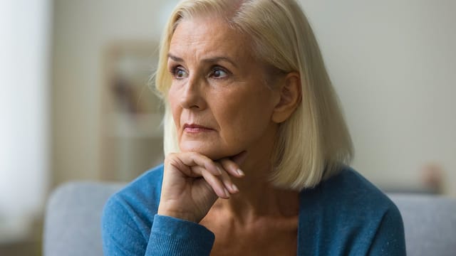 An older woman with short blonde hair sits indoors, resting her chin on her hand and gazing thoughtfully to the side, as if reflecting on regret in old age and worrying about the past, much like findings from a Cornell researcher study.