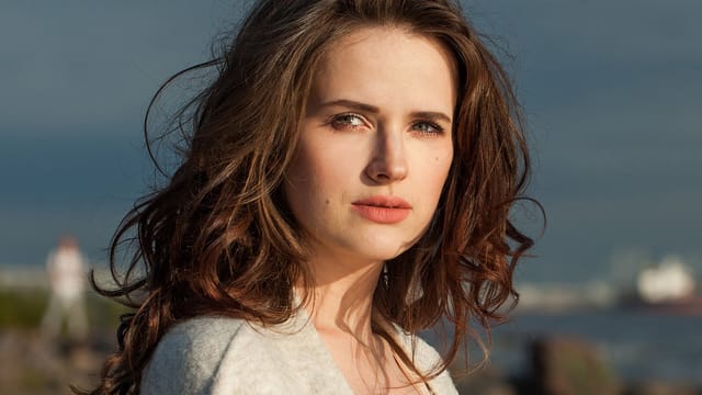 A woman with long, wavy brown hair and light eyes stands outdoors, looking directly at the camera. She wears a light-colored top, her expression subtly reflecting the childhood emotional impact that shapes her presence amid the blurred nature and water.